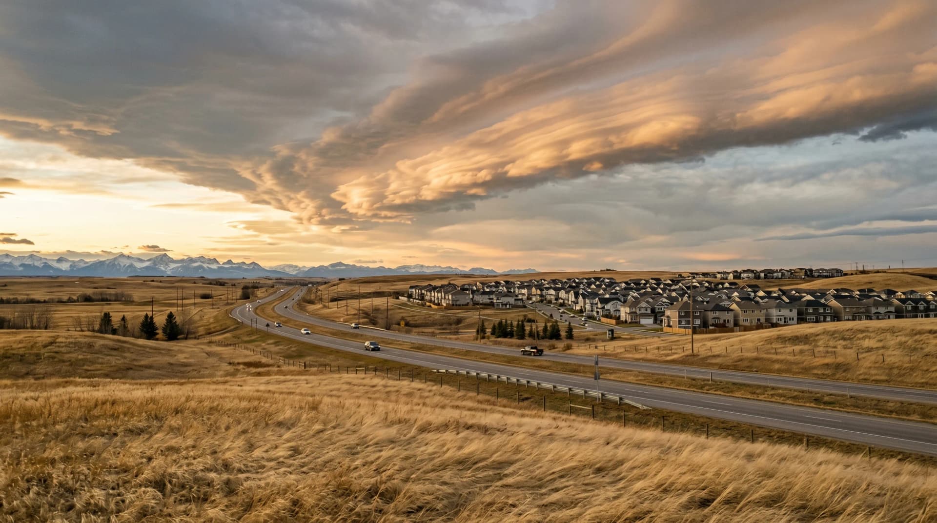 The open Alberta prairie along the QE2 corridor just north of Airdrie at golden hour with a dramatic warm-toned chinook arch overhead, the distant snow-capped Rocky Mountains on the western horizon, a suburban edge of newer two-story homes and golden prairie grass bent low under sustained chinook wind — the Airdrie climate Distinctive Form designs around