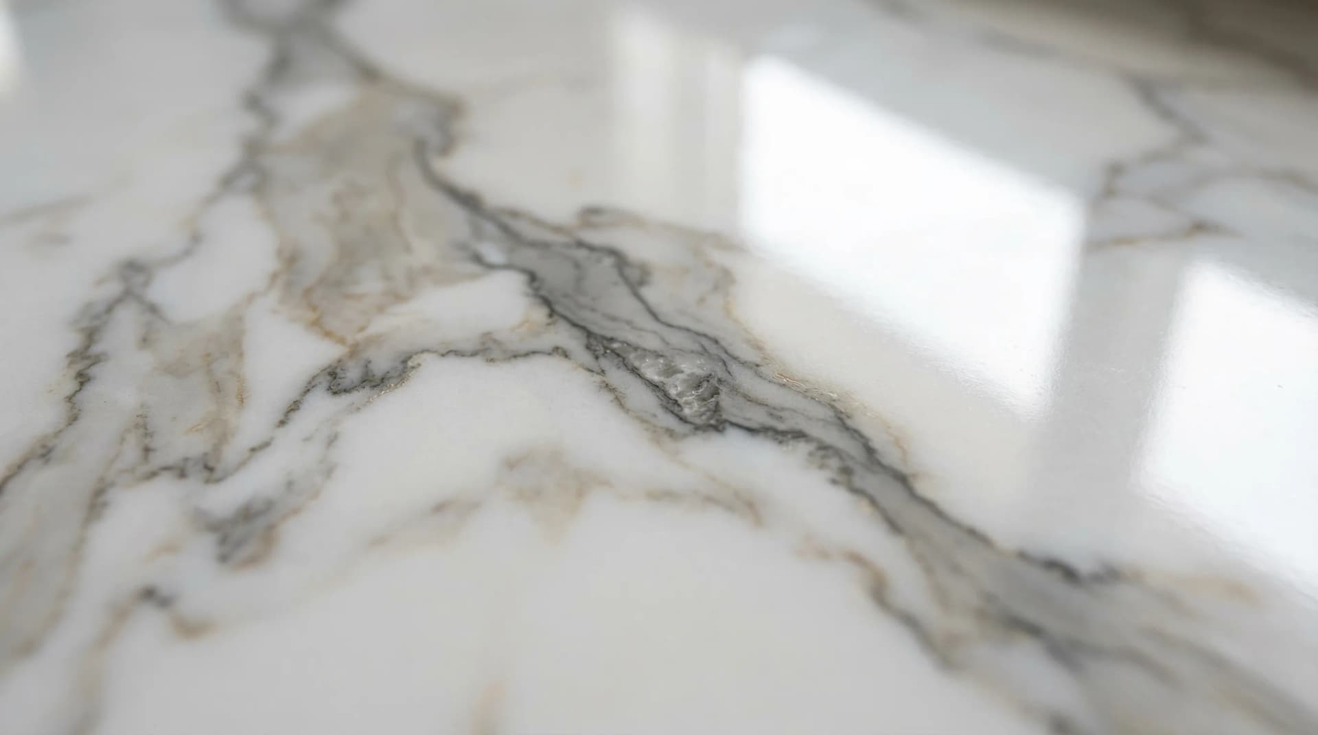 Distinctive Form — premium porcelain surface realistic marble veining macro detail. Extreme close-up of a polished porcelain countertop surface at an oblique angle showing remarkably realistic marble veining — soft grey and warm taupe veins flowing through a luminous white base with fine micro-details mimicking natural calcite crystal formations, the polished surface catching a long streak of reflected light — demonstrating how multi-layer digital decoration on sintered porcelain creates patterns nearly indistinguishable from natural marble.