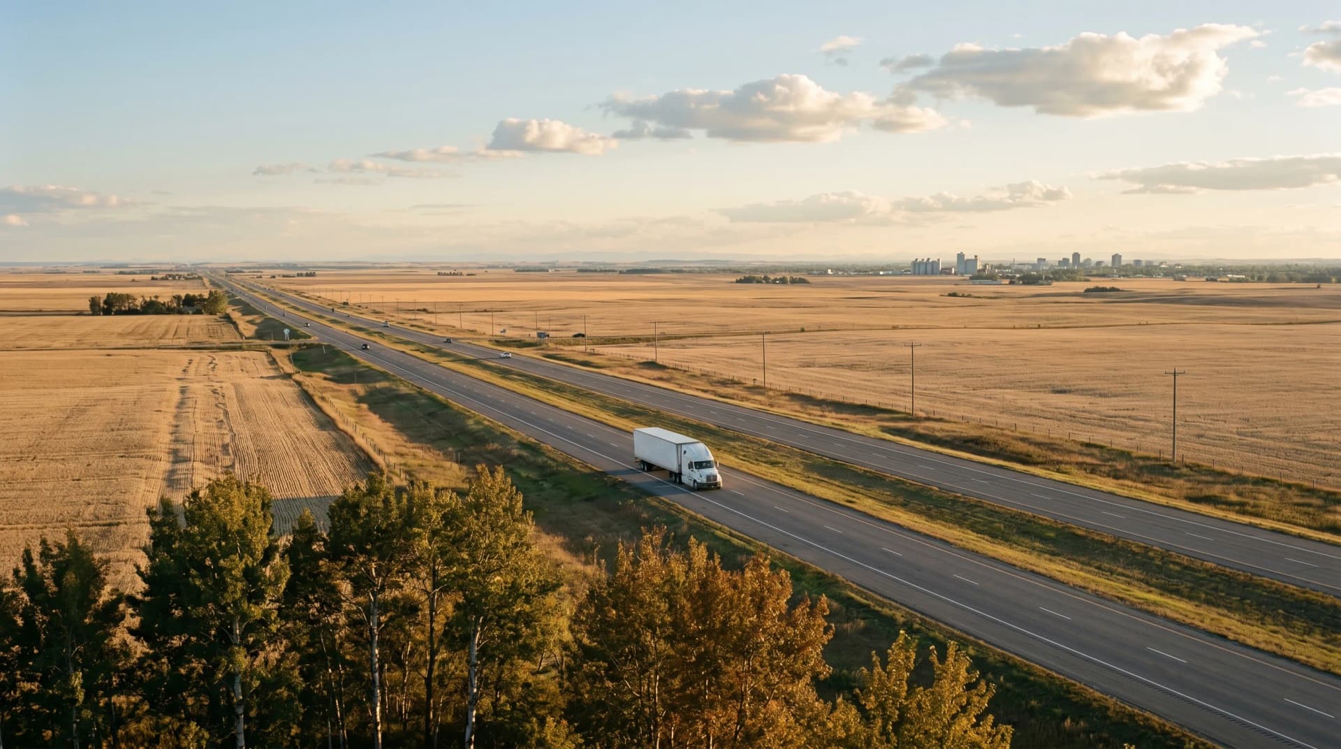 The QE2 Highway corridor cutting across the wide Central Alberta farmland at golden hour with a single transport truck heading south and the distant Red Deer skyline on the horizon — the supply distance Distinctive Form bridges for Red Deer