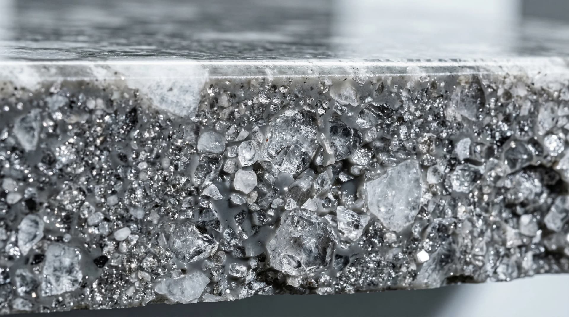 Distinctive Form — engineered quartz composition cross-section. Extreme close-up of an engineered quartz countertop cross-section revealing the internal material structure: individual natural quartz crystal particles of varying sizes — fine glittering powder, medium translucent granules, and larger crystalline chunks — all permanently bonded within a smooth polymer resin matrix, demonstrating the 90–94% quartz mineral content that gives engineered quartz its Mohs 7 hardness rating and non-porous surface properties.