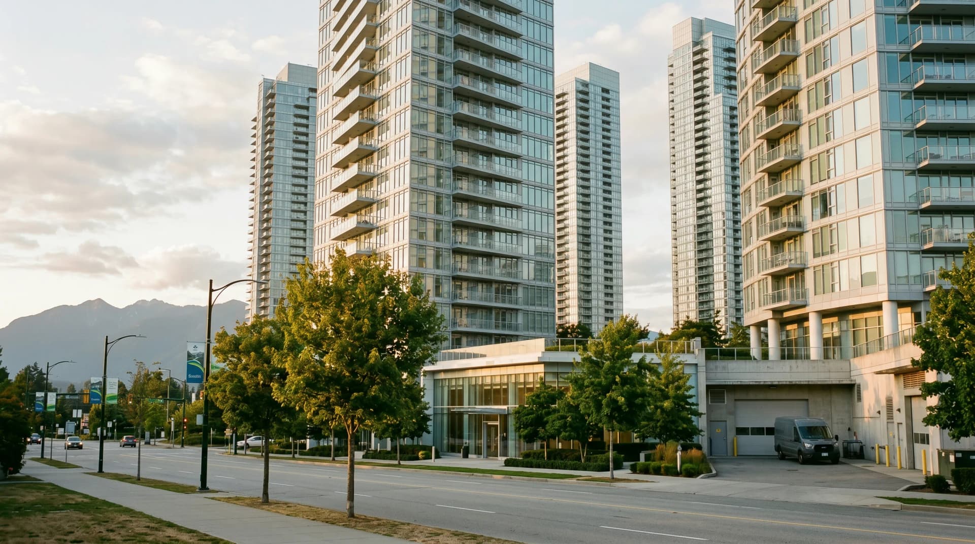 The Brentwood or Metrotown Burnaby high-rise condo cluster with silver glass-and-steel residential towers above a glass entrance lobby and a service loading dock, with the North Shore mountains faintly visible — Distinctive Form serves Burnaby