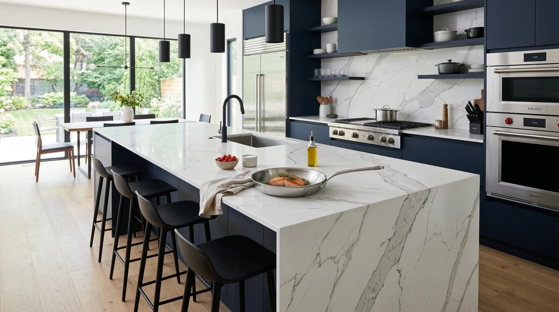 Distinctive Form — modern kitchen with porcelain countertops demonstrating heat resistance. A stunning contemporary kitchen featuring Statuario marble-look porcelain countertops with dramatic grey veining on a large waterfall island with sleek 12 mm thin profile, a stainless steel pan placed directly on the surface demonstrating the material's 316°C heat resistance, dark navy cabinetry, minimalist pendant lighting, and floor-to-ceiling windows — showcasing porcelain's combination of ultra-modern aesthetics and practical performance.
